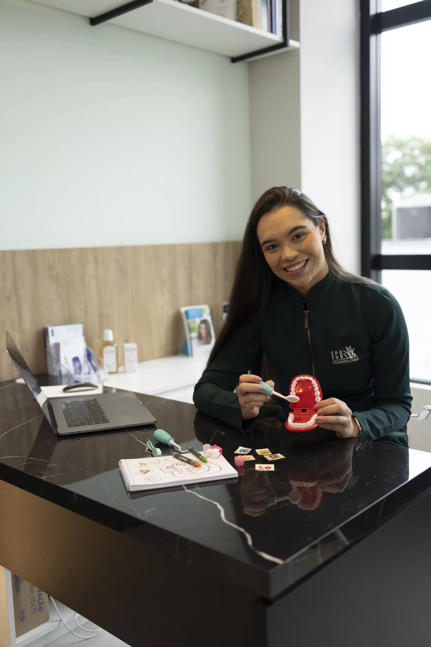 A Dra. Isabel Ribeiro, dentista especializada em odontopediatria na Eco Odontologia, aparece sorridente em seu consultório moderno, apresentando técnicas de escovação de forma lúdica. Sobre a mesa de atendimento, ela utiliza um modelo anatômico de arcada dentária infantil, escova colorida, adesivos e materiais educativos para interagir com as crianças. A imagem enaltece o cuidado atencioso e o ambiente acolhedor preparado pela Dra. Isabel, focando em transformar a consulta em uma experiência leve, positiva e pedagógica, garantindo que os pequenos pacientes desenvolvam hábitos saudáveis com confiança e alegria.