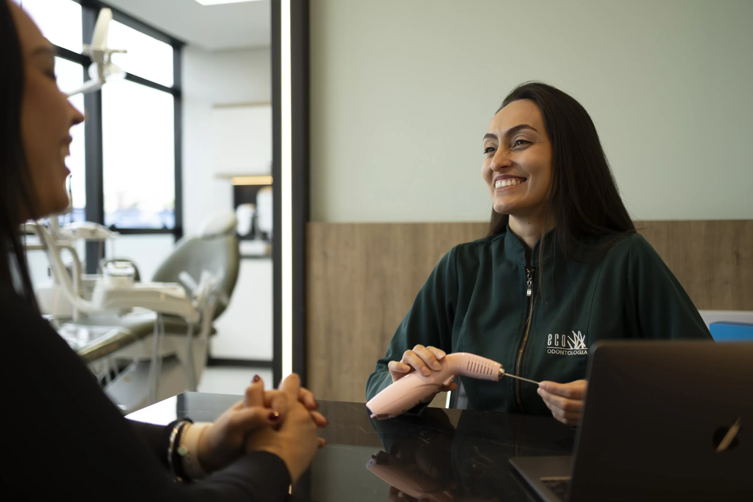 Na clínica Eco Odontologia, uma dentista especializada realiza uma consulta de periodontia, conversando de forma acolhedora e sorridente com uma paciente para planejar o tratamento de saúde gengival. A profissional apresenta equipamentos modernos e explica, com o auxílio de recursos digitais, a importância de prevenir problemas como a gengivite e a periodontite para a longevidade do sorriso. A imagem, capturada no Eco Medical Center em Curitiba, destaca o atendimento personalizado e humanizado da equipe, reforçando o compromisso da clínica em preservar os tecidos de suporte dos dentes com tecnologia avançada, segurança e máximo conforto.