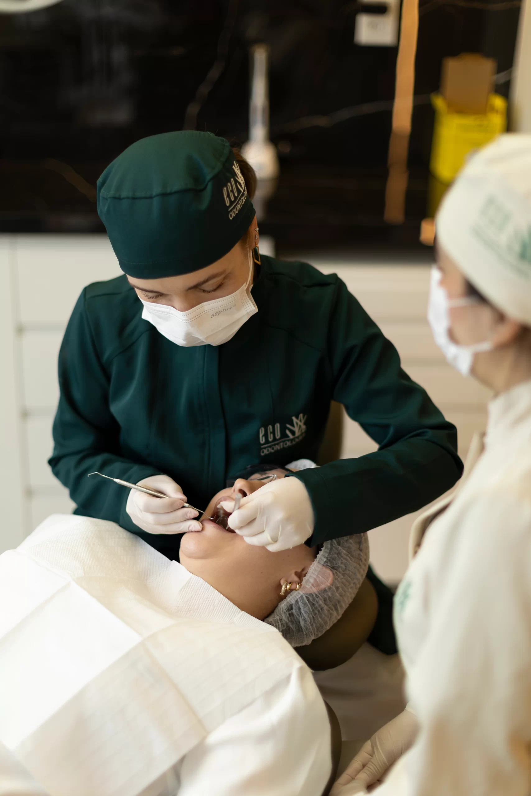 Em um momento de cuidado minucioso na clínica Eco Odontologia, uma dentista especializada realiza a prova de cor e textura para um tratamento de prótese dentária. Através do reflexo de um espelho, a paciente acompanha a seleção precisa das tonalidades, garantindo que a reabilitação oral resulte em um sorriso natural e harmonioso. A imagem destaca o uso de guias de cores profissionais e o atendimento personalizado, evidenciando o compromisso da clínica em aliar tecnologia avançada e sensibilidade estética para restaurar a funcionalidade e a autoestima de cada paciente com máxima excelência.