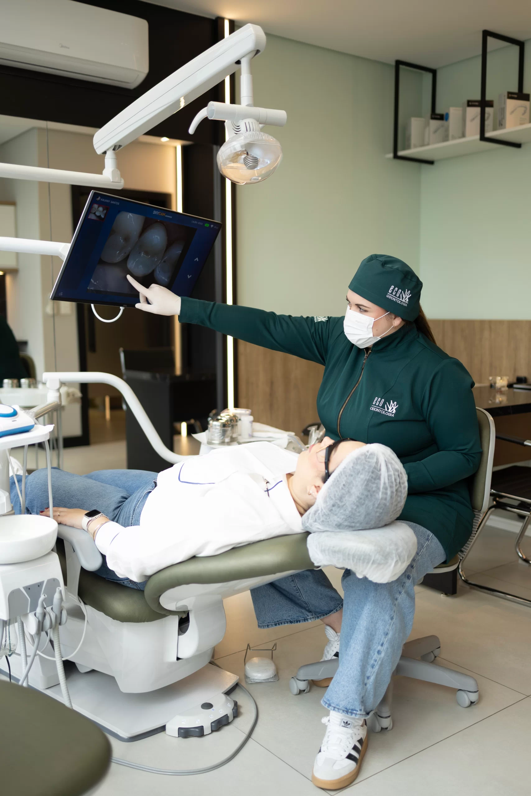Na moderna clínica Eco Odontologia, uma dentista especializada apresenta imagens detalhadas da arcada dentária em um monitor de alta resolução para uma paciente confortavelmente acomodada. A cena no consultório claro e bem equipado destaca a tecnologia de ponta, o atendimento humanizado e o cuidado de excelência que a clínica oferece, garantindo diagnósticos precisos e a melhor experiência em tratamentos de saúde bucal.