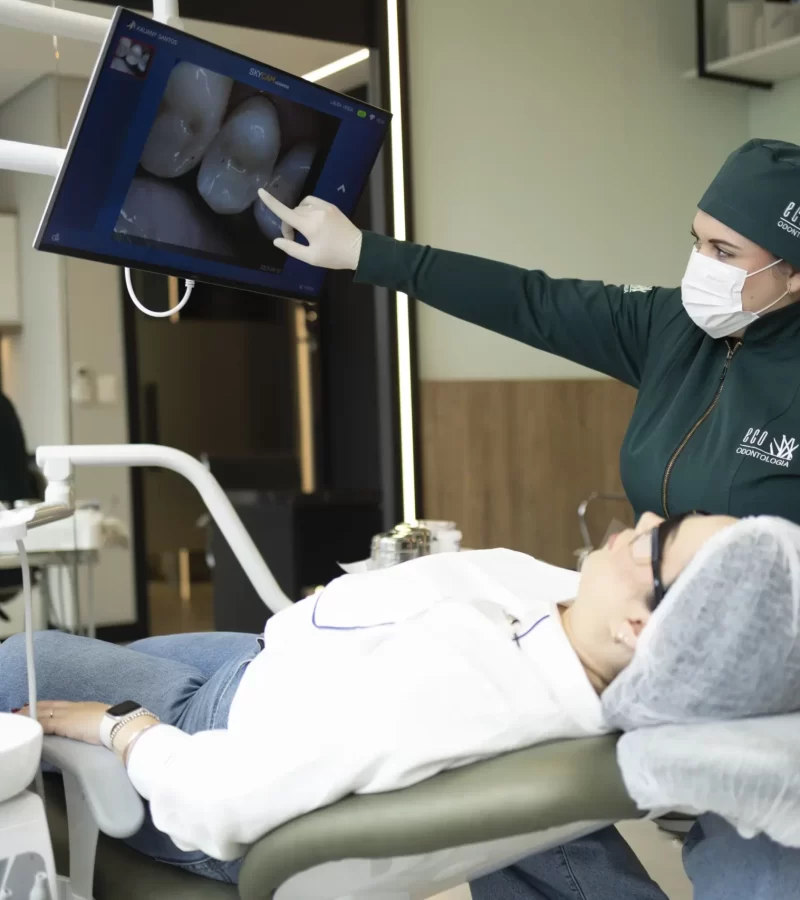Na moderna clínica Eco Odontologia, uma dentista especializada apresenta imagens detalhadas da arcada dentária em um monitor de alta resolução para uma paciente confortavelmente acomodada. A cena no consultório claro e bem equipado destaca a tecnologia de ponta, o atendimento humanizado e o cuidado de excelência que a clínica oferece, garantindo diagnósticos precisos e a melhor experiência em tratamentos de saúde bucal.