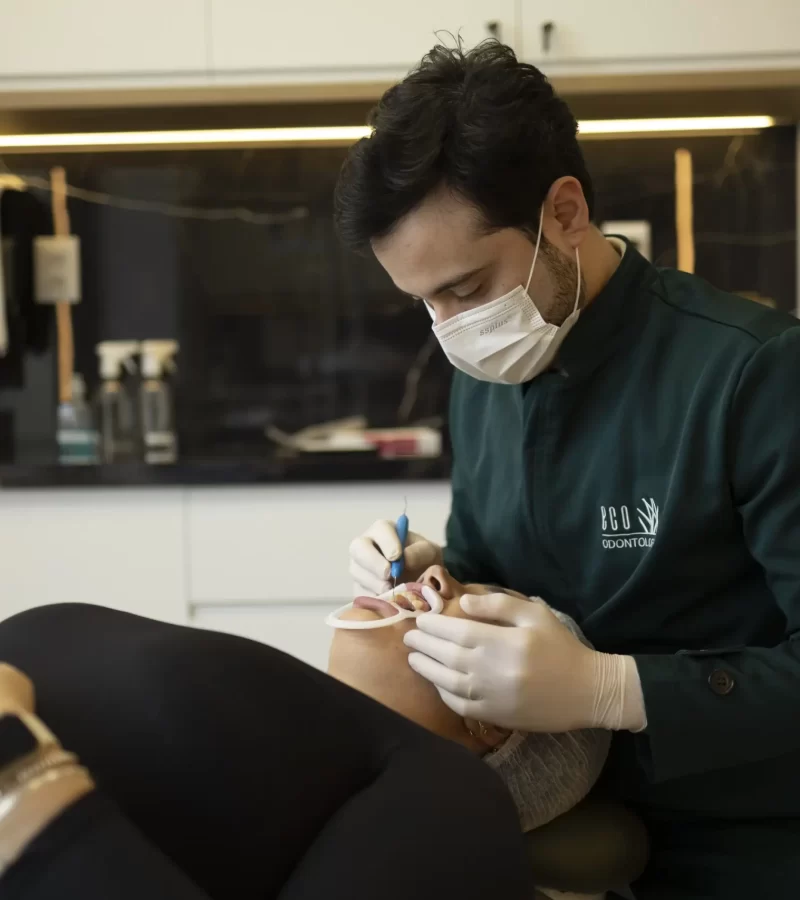 Na moderna clínica Eco Odontologia, um dentista especializado, devidamente paramentado com máscara e luvas, realiza um procedimento de tratamento bucal com precisão em uma paciente confortavelmente deitada na cadeira, que utiliza um afastador labial. A imagem destaca o cuidado, a higiene rigorosa e o profissionalismo da equipe, reforçando o compromisso da clínica com a saúde bucal e o bem-estar de cada paciente durante os atendimentos.