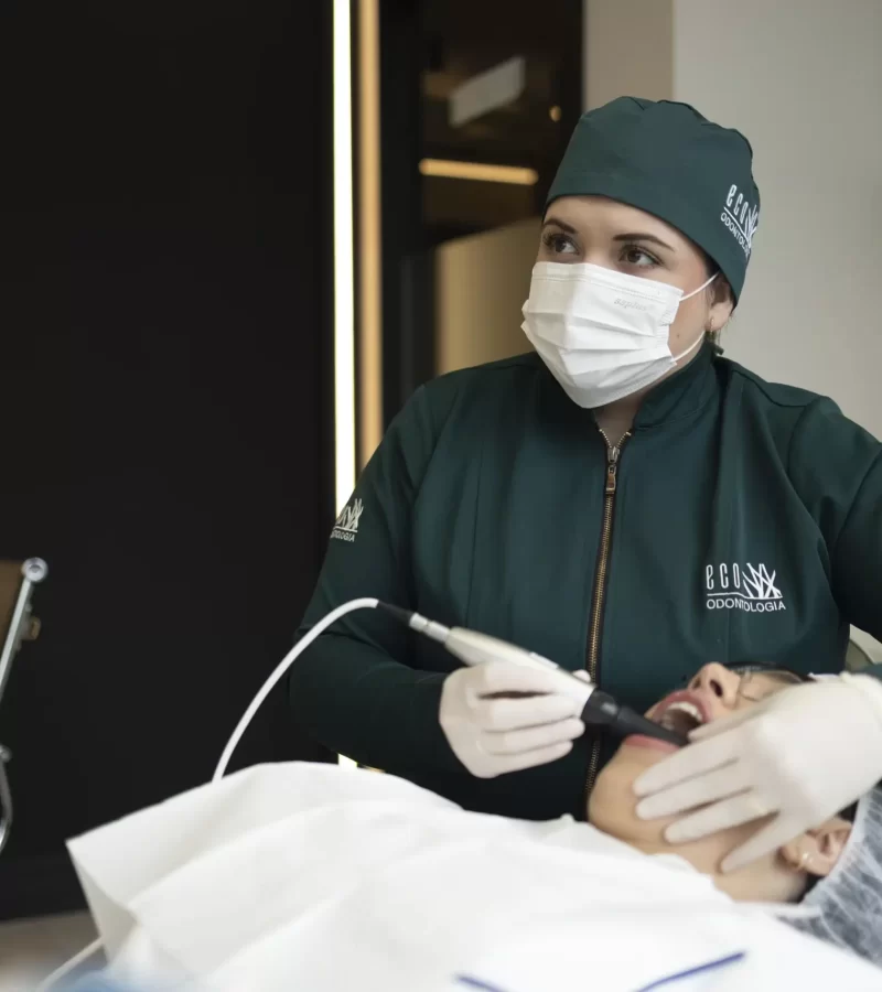Uma dentista da clínica Eco Odontologia utiliza um moderno scanner intraoral digital para mapear a arcada dentária de uma paciente deitada na cadeira do consultório. A cena ilustra o emprego de equipamentos de última geração que modernizam os tratamentos, proporcionando um atendimento odontológico mais ágil, indolor, extremamente confortável e com diagnósticos de altíssima precisão para resultados de excelência.