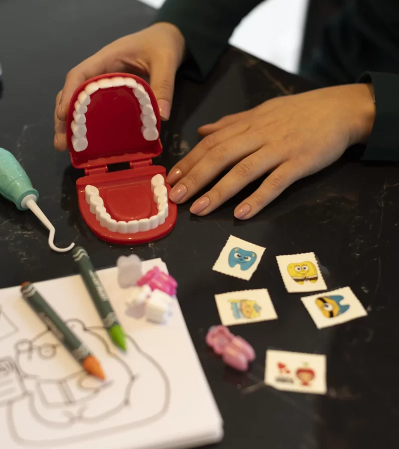 Em um momento de cuidado e educação em saúde bucal na Eco Odontologia, uma profissional utiliza um modelo anatômico de arcada dentária infantil e materiais lúdicos, como adesivos de personagens e desenhos para colorir, para ensinar a escovação correta de forma divertida. A imagem destaca a abordagem acolhedora e pedagógica da odontopediatria na clínica, criando um ambiente leve e positivo para que as crianças aprendam a cuidar do sorriso desde cedo com confiança e sem traumas.