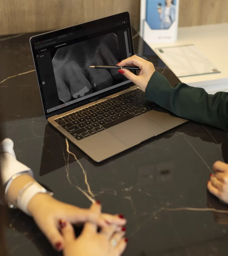 Em uma consulta de planejamento na clínica Eco Odontologia, uma dentista especializada utiliza uma caneta para apontar detalhes em um exame de radiografia digital exibido em um notebook sobre uma elegante mesa de mármore. A imagem destaca a precisão diagnóstica e a transparência no atendimento, onde a tecnologia é utilizada para explicar cada etapa do tratamento de forma clara ao paciente. A cena reforça o compromisso da clínica com a excelência técnica e o uso de recursos digitais avançados para garantir os melhores resultados em saúde e estética bucal.