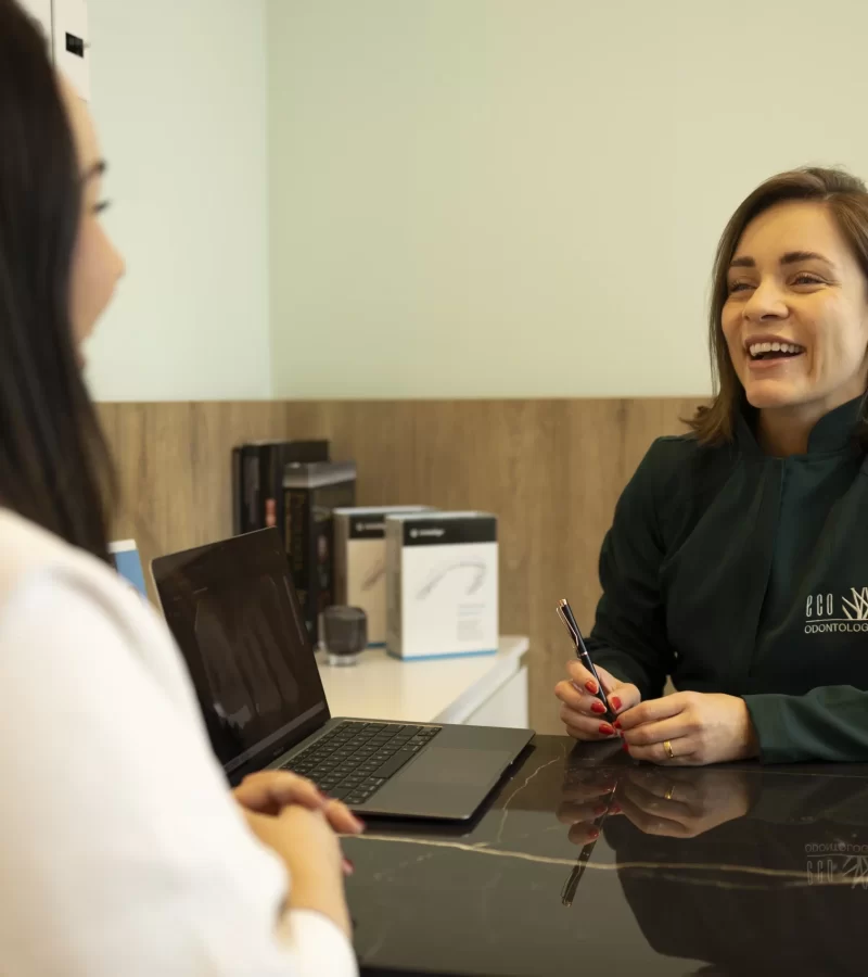 Na moderna clínica Eco Odontologia, uma dentista acolhedora e sorridente conversa amigavelmente com uma paciente durante uma consulta de avaliação. Sentadas em uma sala confortável e sofisticada, com um notebook exibindo exames digitais sobre a mesa, a cena transmite confiança, transparência e um atendimento focado na humanização. A imagem destaca o cuidado e a dedicação da equipe em oferecer um planejamento odontológico totalmente personalizado, garantindo a melhor experiência, clareza nos diagnósticos e excelência na saúde bucal.