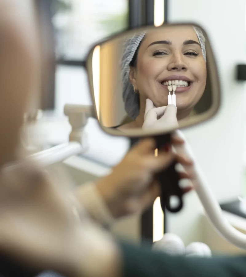 Através de um espelho de mão, uma paciente sorridente acompanha o processo de seleção da cor ideal para seu tratamento estético na Eco Odontologia. Uma profissional especializada utiliza uma escala de cores técnica (Vita Shade Guide) para comparar diferentes tonalidades diretamente com os dentes da paciente, garantindo um resultado natural e personalizado. A imagem captura o cuidado minucioso com a estética e a satisfação do paciente, destacando o compromisso da clínica em transformar sorrisos com precisão técnica e atenção aos desejos individuais.